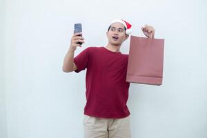 giovane asiatico uomo indossare un' Santa Claus cappello Tenere un' smartphone e un' shopping Borsa con espressioni di Sorridi, shock, e sorpresa, isolato contro un' bianca sfondo per visivo comunicazione foto