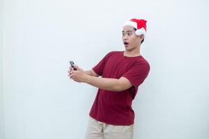 giovane asiatico uomo indossare un' Santa Claus cappello Tenere un' smartphone e esprimendo un' Sorridi, shock e sorpresa isolato di un' bianca sfondo per visivo comunicazione foto