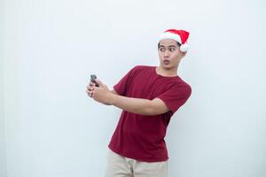 giovane asiatico uomo indossare un' Santa Claus cappello Tenere un' smartphone e esprimendo un' Sorridi, shock e sorpresa isolato di un' bianca sfondo per visivo comunicazione foto