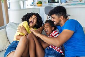 allegro persone seduta su divano nel vivente camera avere divertimento poco figlia solletico madre ridendo insieme con genitori godere gratuito tempo giocando a casa. fine settimana attività contento famiglia stile di vita concetto foto