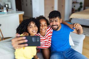 contento africano coppia di genitori e ridendo figlia riposo su divano Tenere smartphone, assunzione autoscatto su cellulare, fabbricazione video chiamata, guardare a mobile Telefono schermo, sorridente, ridacchiando. foto