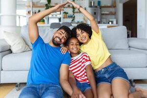 nuovo edificio Residenziale Casa Acquista appartamento concetto. elegante pieno famiglia con uno ragazzo seduta su tappeto, mamma e papà con figlia fabbricazione tetto figura con mani braccia al di sopra di teste foto