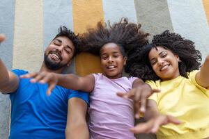 ritratto di contento famiglia in posa su pavimento a casa, posa su tappeto e legame, allegramente sorridente, Visualizza sopra. positivo padre, madre e figlia coccole, superiore Visualizza foto