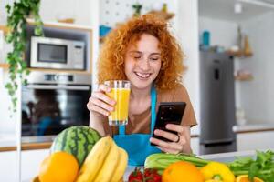 sorridente bella donna guardare a mobile Telefono e Tenere bicchiere di arancia succo mentre cucinando fresco verdure nel cucina interno a casa foto