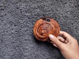 mano Tenere cromboloni, il giro brioche, nuovo York rotolo, un' virale Pasticcino combinando Cornetti e bomboloni su calcestruzzo parete sfondo. foto