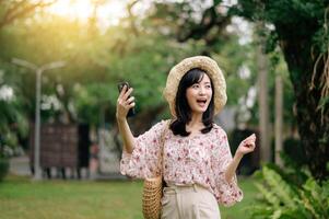 ritratto di asiatico giovane donna viaggiatore con tessitura cappello e cestino utilizzando mobile Telefono su verde parco natura sfondo. viaggio viaggio stile di vita, mondo viaggio esploratore o Asia estate turismo concetto. foto