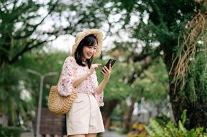 ritratto di asiatico giovane donna viaggiatore con tessitura cappello e cestino utilizzando mobile Telefono su verde parco natura sfondo. viaggio viaggio stile di vita, mondo viaggio esploratore o Asia estate turismo concetto. foto