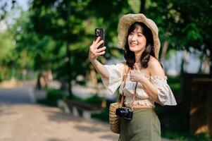 ritratto di asiatico giovane donna viaggiatore con tessitura cappello, cestino, mobile Telefono e telecamera su verde pubblico parco sfondo. viaggio viaggio stile di vita, mondo viaggio esploratore o Asia estate turismo concetto. foto