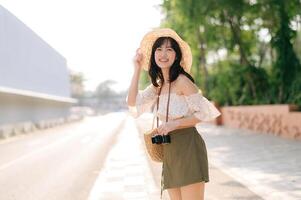 ritratto di asiatico giovane donna viaggiatore con tessitura cappello e cestino e un' telecamera in piedi di il strada. viaggio viaggio stile di vita, mondo viaggio esploratore o Asia estate turismo concetto. foto