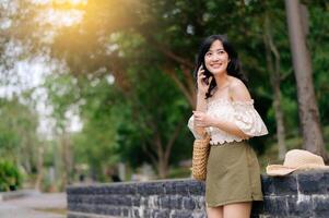 ritratto di asiatico giovane donna viaggiatore con tessitura cappello, cestino, mobile Telefono e telecamera su verde pubblico parco sfondo. viaggio viaggio stile di vita, mondo viaggio esploratore o Asia estate turismo concetto. foto