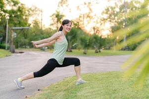 femmina pareggiatore. in forma giovane asiatico donna con verde abbigliamento sportivo allungamento muscolo nel parco prima in esecuzione e godendo un' salutare all'aperto. fitness corridore ragazza nel pubblico parco. benessere essere concetto foto