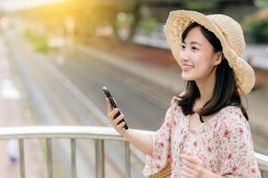 giovane asiatico donna viaggiatore con tessitura cestino utilizzando mobile Telefono e in piedi su cavalcavia con ferrovia sfondo. viaggio viaggio stile di vita, mondo viaggio esploratore o Asia estate turismo concetto. foto