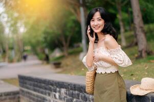 ritratto di asiatico giovane donna viaggiatore con tessitura cappello, cestino, mobile Telefono e telecamera su verde pubblico parco sfondo. viaggio viaggio stile di vita, mondo viaggio esploratore o Asia estate turismo concetto. foto