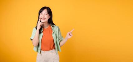 dentale Salute con travolgente giovane asiatico donna anni '30, indossare arancia camicia e verde Maglione punti per sua denti, isolato su vivace giallo sfondo, sottolineando il importanza di dentale assistenza sanitaria. foto