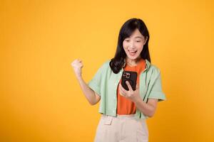 il gioia di grande notizia come un' giovane asiatico donna nel sua anni '30, indossare arancia camicia e verde Maglione, celebra con un' cazzotto su gesto e smartphone su giallo studio sfondo. mobile concetto. foto