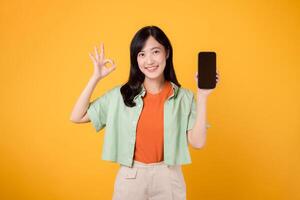 eccitazione di un' nuovo mobile applicazione con un' allegro giovane asiatico donna anni '30, indossare arancia camicia e verde Maglione, visualizzazione smartphone schermo con va bene mano gesto su giallo studio sfondo. foto