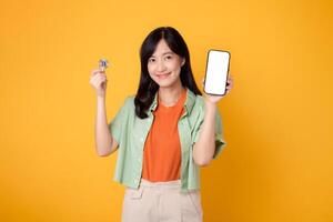 giovane 30s asiatico donna contento viso vestito nel arancia camicia e verde Maglione mostrando smartphone schermo Schermo e crypto moneta moneta isolato su giallo sfondo. futuro finanza concetto. foto
