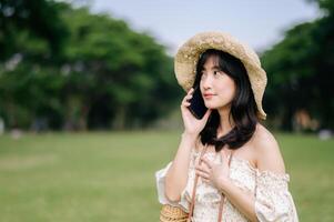 ritratto di asiatico giovane donna viaggiatore con tessitura cappello, cestino, mobile Telefono e telecamera su verde pubblico parco sfondo. viaggio viaggio stile di vita, mondo viaggio esploratore o Asia estate turismo concetto. foto