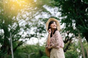 ritratto di asiatico giovane donna viaggiatore con tessitura cappello e cestino contento Sorridi su verde pubblico parco natura sfondo. viaggio viaggio stile di vita, mondo viaggio esploratore o Asia estate turismo concetto. foto