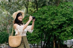 ritratto di asiatico giovane donna viaggiatore con tessitura cappello e cestino e un' telecamera su verde pubblico parco natura sfondo. viaggio viaggio stile di vita, mondo viaggio esploratore o Asia estate turismo concetto. foto