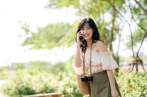 ritratto di asiatico giovane donna viaggiatore con tessitura cappello, cestino, mobile Telefono e telecamera su verde pubblico parco sfondo. viaggio viaggio stile di vita, mondo viaggio esploratore o Asia estate turismo concetto. foto