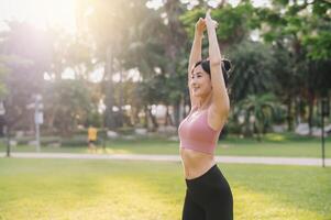 benessere e salutare stile di vita ritratto di 30s asiatico donna nel rosa abbigliamento sportivo. preparare e allungare braccio muscoli prima tramonto correre nel il parco. fitness al di fuori e vivere un' equilibrato vita. foto