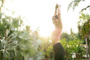 in forma 30s giovane asiatico donna nel rosa abbigliamento sportivo giochi yoga e tratti muscoli. abbraccio salutare all'aperto stile di vita con fitness yoga ragazza nel pubblico parco a tramonto. benessere e bene essere concetto. foto