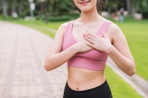 donna pareggiatore. giovane asiatico femmina contento Sorridi indossare rosa abbigliamento sportivo Tenere mani su il petto prima in esecuzione nel pubblico parco. assistenza sanitaria benessere concetto. foto