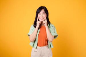 dentale cura con giovane asiatico donna anni '30, vestito nel arancia camicia e verde Maglione punti per sua denti, isolato su un' vivace giallo sfondo, evidenziazione il importanza di dentale assistenza sanitaria foto