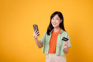 shopping a partire dal mobile con un' travolgente giovane asiatico donna nel sua anni '30, elegantemente vestito nel arancia camicia e verde Maglione, utilizzando smartphone mentre Tenere credito carta su giallo studio sfondo. foto