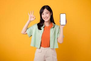 il innovazione di nuovo mobile applicazione con giovane asiatico donna nel sua anni '30, vestito nel arancia camicia e verde Maglione, presentazione smartphone schermo con va bene mano gesto su giallo studio sfondo. foto