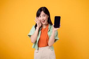 giovane asiatico donna nel sua anni '30, elegantemente vestito nel arancia camicia e verde Maglione, utilizzando smartphone schermo con un' grido bocca su giallo studio sfondo. nuovo mobile dispositivo e in linea shopping concetto. foto