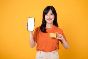 entusiasta asiatico donna nel sua anni '30, in mostra vuoto smartphone schermo e credito carta su vivace giallo sfondo. veloce in linea pagamento acquisti. foto