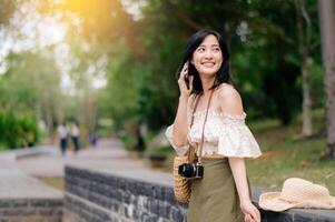 ritratto di asiatico giovane donna viaggiatore con tessitura cappello, cestino, mobile Telefono e telecamera su verde pubblico parco sfondo. viaggio viaggio stile di vita, mondo viaggio esploratore o Asia estate turismo concetto. foto