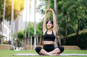 ritratto in forma giovane asiatico donna 30s indossare nero abbigliamento sportivo ascolta rilassante musica mentre allungamento muscolo prima yoga esercizio su yoga stuoia nel verde natura parco. benessere concetto. foto