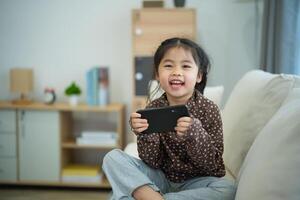 asiatico bambino ragazza guardare utilizzando e toccare mobile Telefono schermo su divano divano. bambino sorridente divertente tempo per uso mobile Telefono. pure tanto schermo volta. carino ragazza Guardando video mentre tv, Internet dipendenza. foto