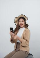 asiatico sorridente turista donna con bretelle indossare cappello e seduta bagaglio Tenere mobile Telefono, copia spazio andando per viaggio su vacanze isolato su blu studio sfondo. viaggio viaggio concetto. foto