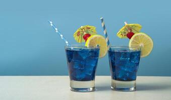 Due blu cocktail nel un' bicchiere pieno con ghiaccio guarnito con Limone fette, ciliegie, giallo ombrelli e sorbetti su un' bianca tavolo foto