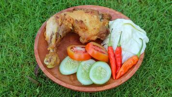 fritte pollo con lalapan, fritte pollo con fresco la verdura, pomodoro fette, cetriolo e Sambal autentico ricetta di indonesiano pollo lalapan. fritte pollo su verde erba sfondo. foto