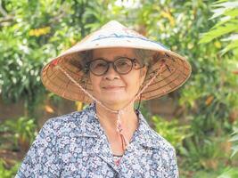 un' ritratto di un asiatico anziano donna indossare cappello, sorridente e guardare a telecamera mentre in piedi nel un' giardino. spazio per testo. concetto di vecchio persone e assistenza sanitaria foto