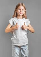 contento bambino, poco ragazza mostrando pollici su gesto nel un' bianca maglietta foto