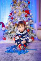 un' gioioso poco ragazzo ammirazione il festivo Natale albero. un' poco ragazzo seduta nel davanti di un' Natale albero foto