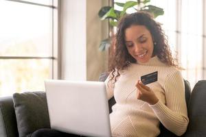 donna latina che utilizza laptop e mano che tiene la carta di credito per lo shopping sul divano foto