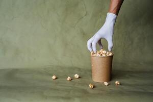 mano nel un' bianca guanto raggiunge per Popcorn, prendere cibo nel protettivo guanti, hold un' pezzo di cibo con Due dita, un' pieno scatola di Popcorn, sicurezza a il veloce cibo distribuzione, minimo concetto foto