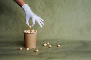 mano nel un' bianca guanto raggiunge per Popcorn, prendere cibo nel protettivo guanti, hold un' pezzo di cibo con Due dita, un' pieno scatola di Popcorn, sicurezza a il veloce cibo distribuzione, minimo concetto foto