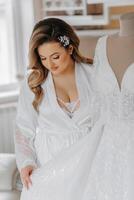 mattina di il sposa prima il nozze. un' bellissimo giovane donna con un' velo e un' bellissimo acconciatura nel un' bianca accappatoio vicino sua nozze vestito su un' manichino. professionale trucco. ultimo preparativi. foto