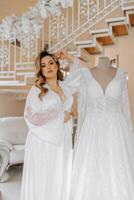 mattina di il sposa prima il nozze. un' bellissimo giovane donna con un' velo e un' bellissimo acconciatura nel un' bianca accappatoio vicino sua nozze vestito su un' manichino. professionale trucco. ultimo preparativi. foto