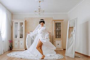 mattina di il sposa prima il nozze. un' bellissimo giovane donna con un' velo e un' bellissimo acconciatura nel un' bianca accappatoio vicino sua nozze vestito su un' manichino. professionale trucco. ultimo preparativi. foto