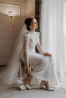 moda ritratto di un' bellissimo sposa nel un' lussuoso nozze vestito con pizzo e cristalli nel un Arabo interno stile. bellissimo sposa mette su scarpe. preparazione per il nozze cerimonia. foto