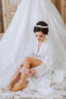 mattina di il sposa prima il nozze. un' bellissimo giovane donna con un' velo e un' bellissimo acconciatura nel un' bianca accappatoio vicino sua nozze vestito su un' manichino. professionale trucco. ultimo preparativi. foto